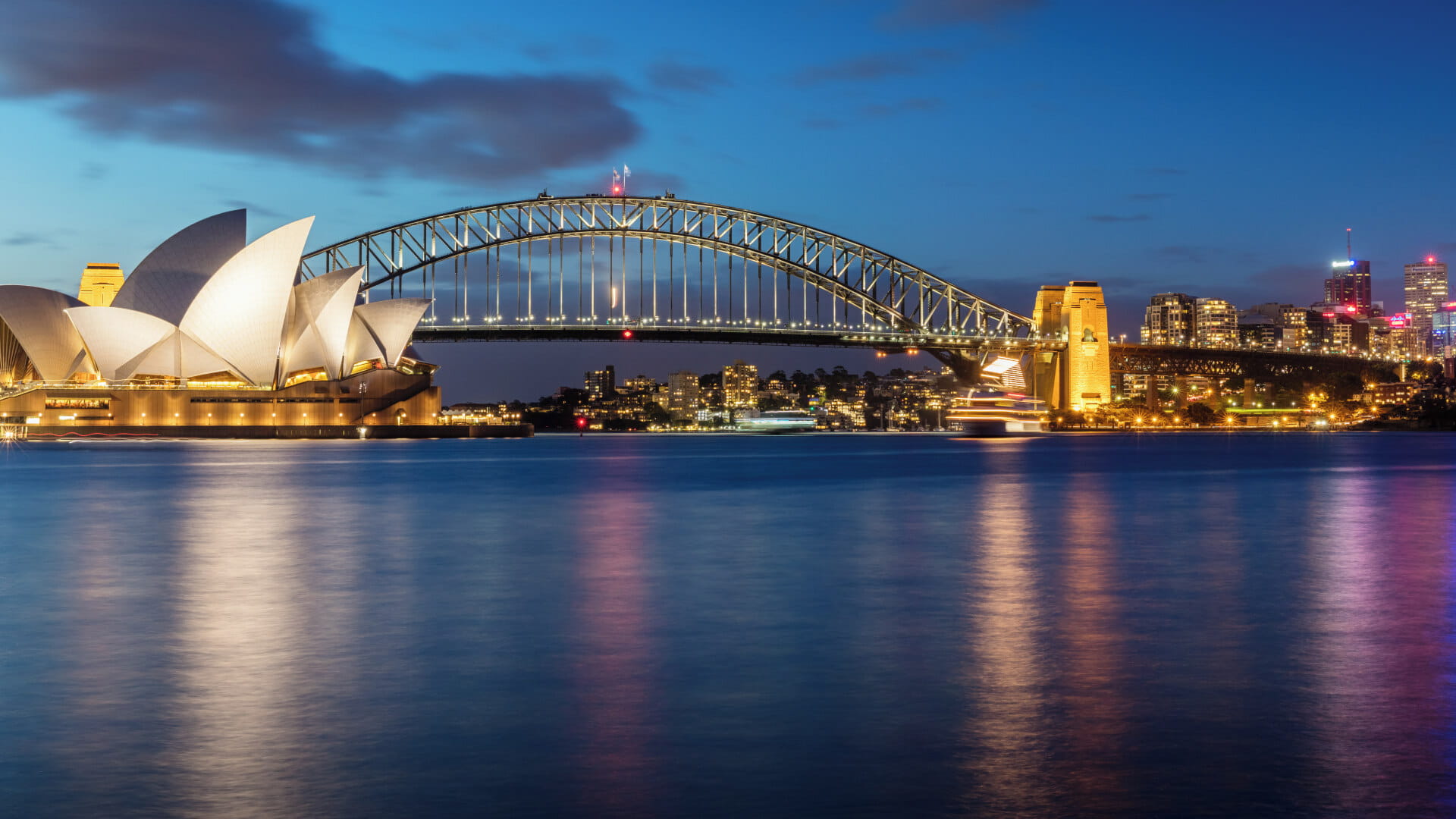 A stunning night view of the iconic Sydney Harbour Bridge arching over the sparkling waters of Sydney Harbour, with the city skyline in the background.